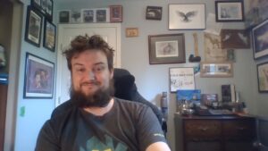 A picture of James in his home office. White male with unkempt beard in grey t-shirt.